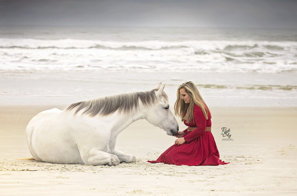 Girl in red dress connecting with horse laying down on beach