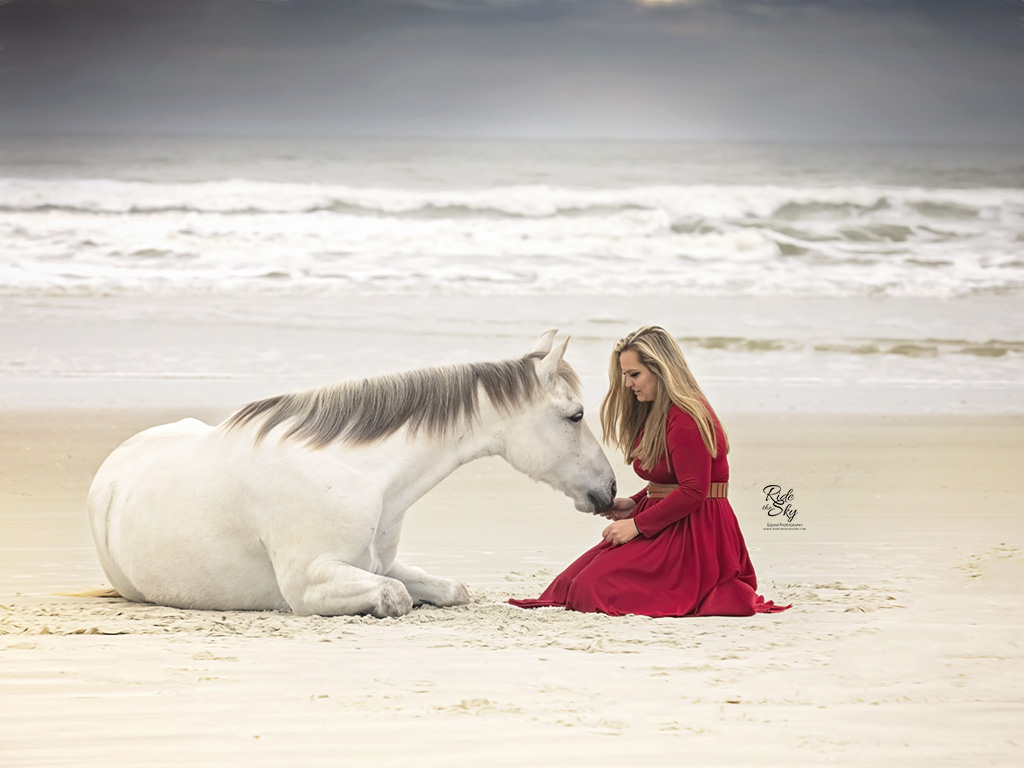 Girl in red dress connecting with horse laying down on beach