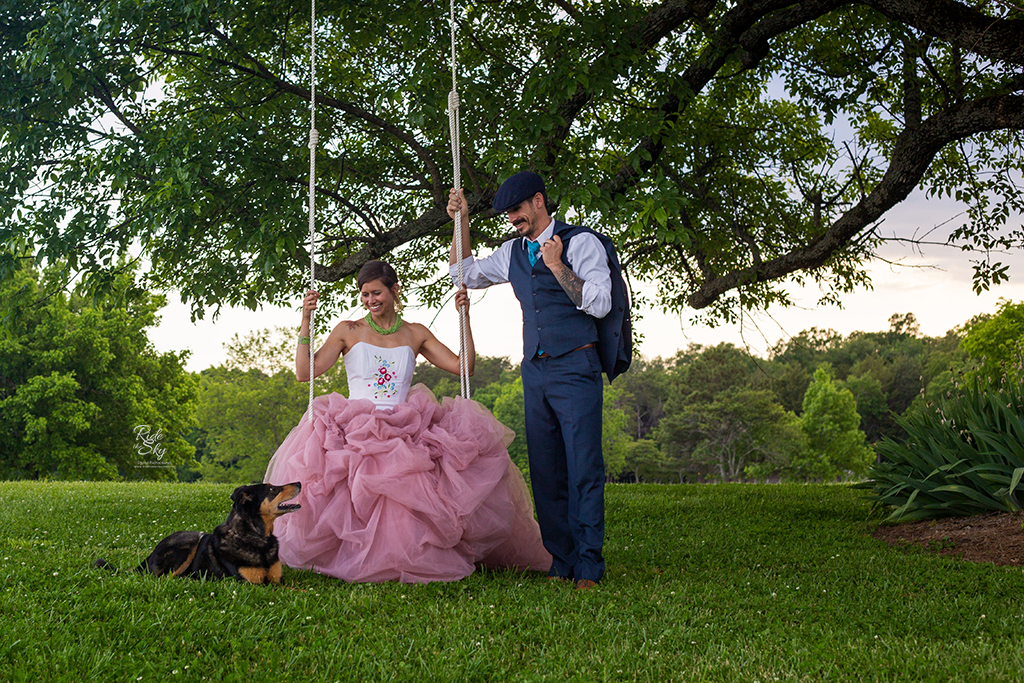 Women in pink tulle skirt on swing with man in suit and dog standing next to her at Kensington Cove Farm in Chickamauga GA