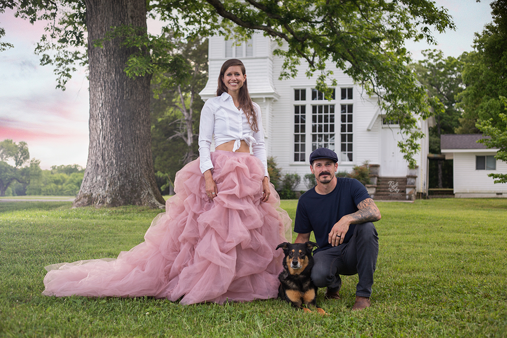 Woman standing in pink skirt with train next to crouching man and dog outside of white chirch and tree and man