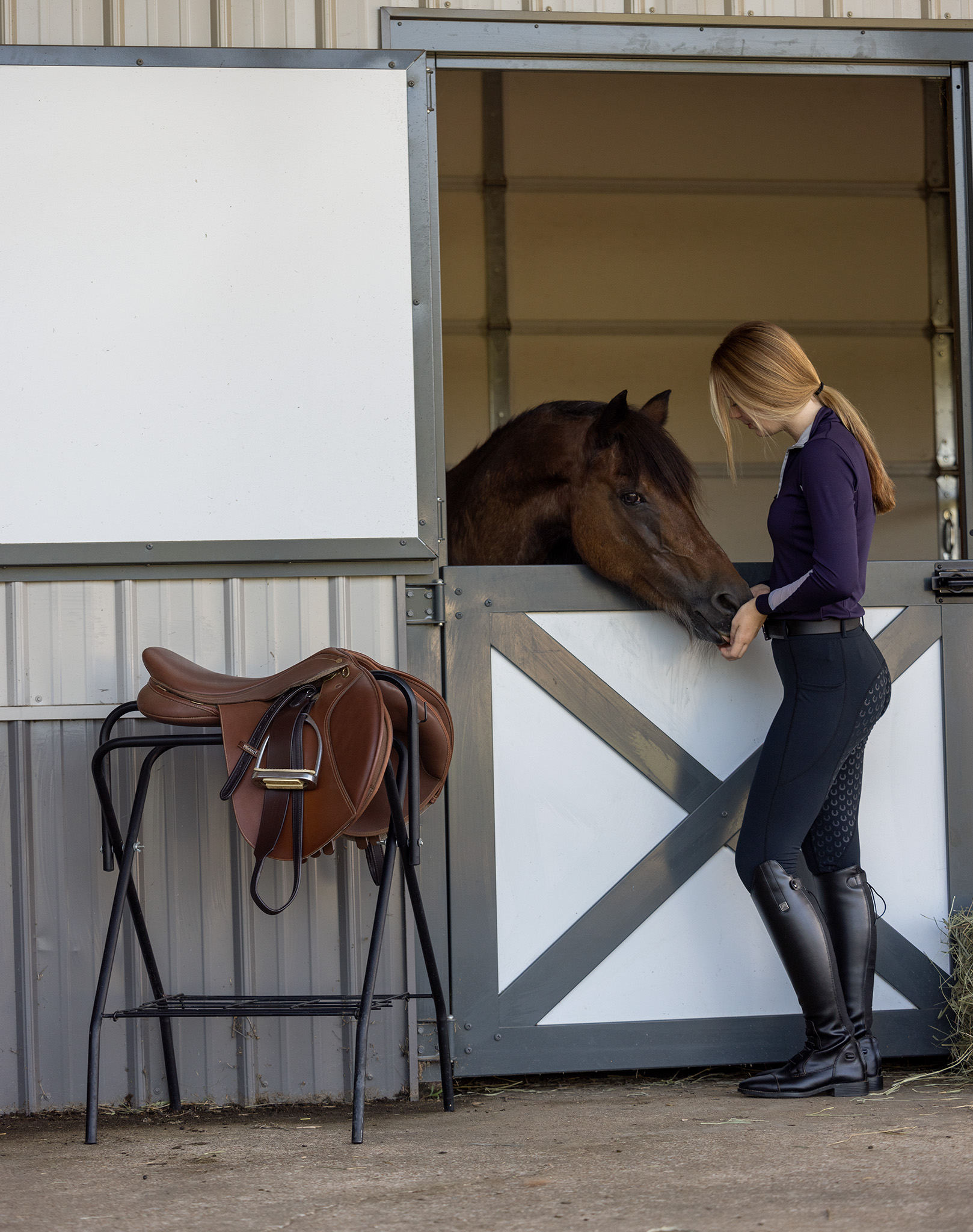 Commercial and branding image of Equestrian girl feeding horse a treat in stable with english saddle and total saddle fit stirrup leathers sitting nearby.